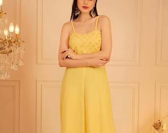 Può includere: Tuta gialla con corpetto con perline e pantaloni a gamba larga. La parte superiore presenta spalline sottili e un design dettagliato. I pantaloni sono in tessuto liscio e fluido. L'outfit è di un colore brillante e uniforme.