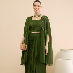 May include: An olive green outfit comprising a crop top, draped skirt, and cape, adorned with silver sequins. The ensemble is accessorized with a gold clutch, necklace, and sandals. The background is a neutral beige colour.