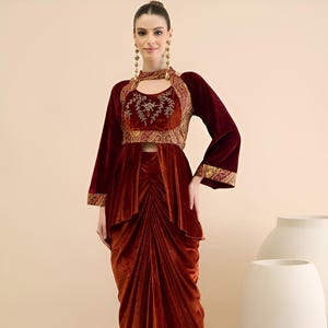 May include: A burgundy velvet outfit comprising a crop top with gold embroidery and long sleeves, and a draped skirt. The ensemble includes a matching choker and gold earrings. The style is elegant and sophisticated, suitable for special occasions.