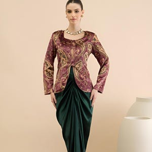 May include: A woman models a two-piece outfit. The top is a burgundy paisley print jacket with long sleeves. The bottom is a dark green, draped skirt. The outfit is paired with gold sandals and a pearl necklace.