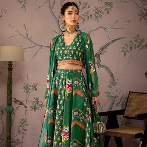 May include: A green floral print outfit comprising a crop top, long skirt, and sheer jacket. The ensemble features a V-neck and is decorated with colourful floral patterns. The skirt is floor-length, and the jacket has long sleeves. The outfit is accessorised with jewellery and a small green purse.