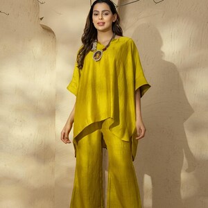 Op de afbeelding: Een tweedelige outfit in een levendige chartreuse kleur. De set bestaat uit een gedrapeerde, asymmetrische top en een broek met wijde pijpen. Een opvallende ketting met een ronde hanger voegt een contrast toe. De outfit is gemaakt van een textuurstof.