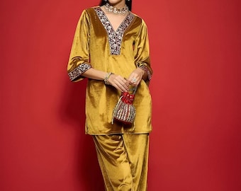 Puede incluir: Un conjunto de terciopelo amarillo mostaza, que consta de una túnica y pantalones a juego. La túnica presenta un cuello en V y adornos decorativos con bordados florales. El conjunto se complementa con joyas y una pequeña bolsa con cuentas. El fondo es rojo liso.