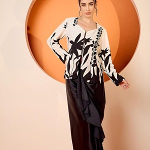 May include: A two-piece outfit featuring a cream jacket with a black floral design and a black skirt. The jacket has a zip closure and long sleeves. The skirt has a draped design. The outfit is suitable for formal occasions.