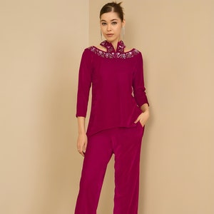 Puede incluir: Un conjunto de dos piezas de terciopelo magenta. La parte superior tiene un escote único con adornos plateados y mangas tres cuartos. Los pantalones a juego son de pierna ancha. La modelo lleva tacones plateados con tiras.