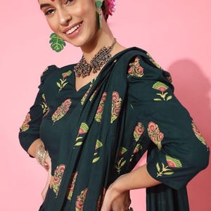 May include: A woman wearing a dark green dress with a floral print and a matching dupatta. The dress has a V-neckline and three-quarter sleeves. She is wearing silver jewelry, including a necklace and earrings.