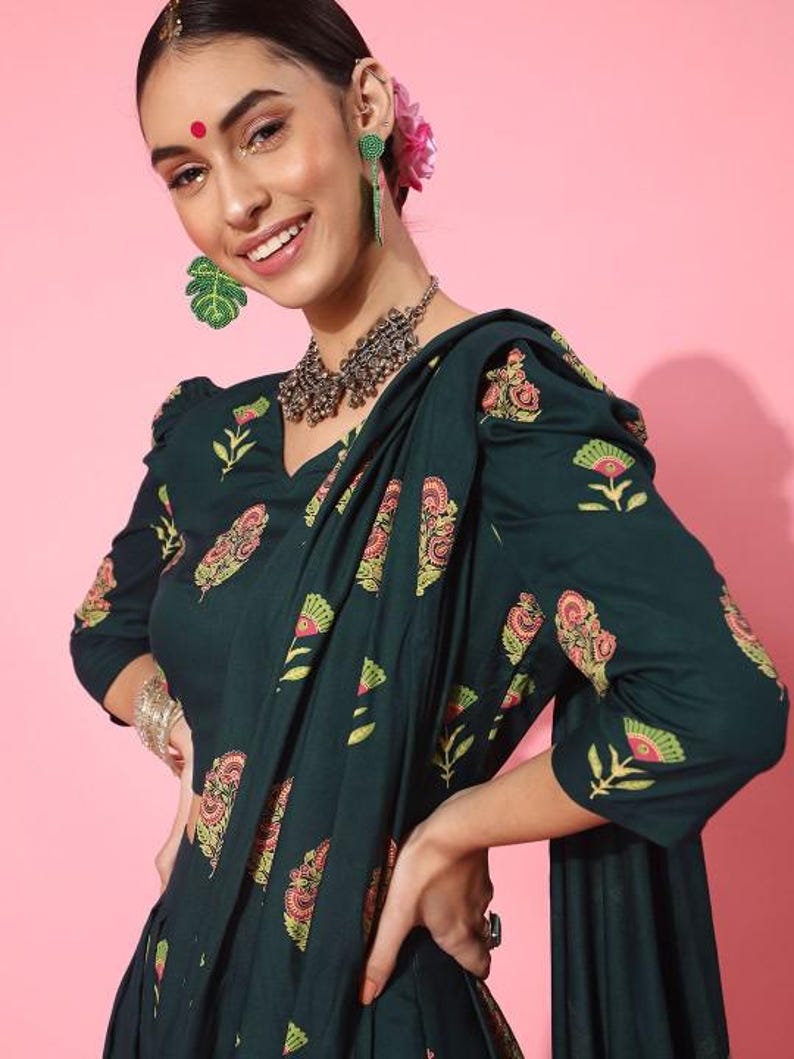 May include: A woman wearing a dark green dress with a floral print and a matching dupatta. The dress has a V-neckline and three-quarter sleeves. She is wearing silver jewelry, including a necklace and earrings.