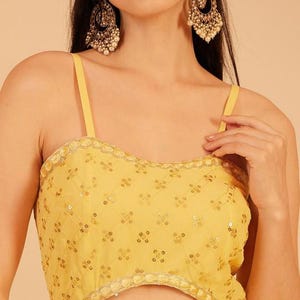 Pode incluir: Vestido de noite amarelo com decote em cora&ccedil;&atilde;o e recorte em forma de fechadura. O vestido &eacute; adornado com lantejoulas e um padr&atilde;o floral. A modelo usa brincos dourados grandes e ornamentados com p&eacute;rolas.