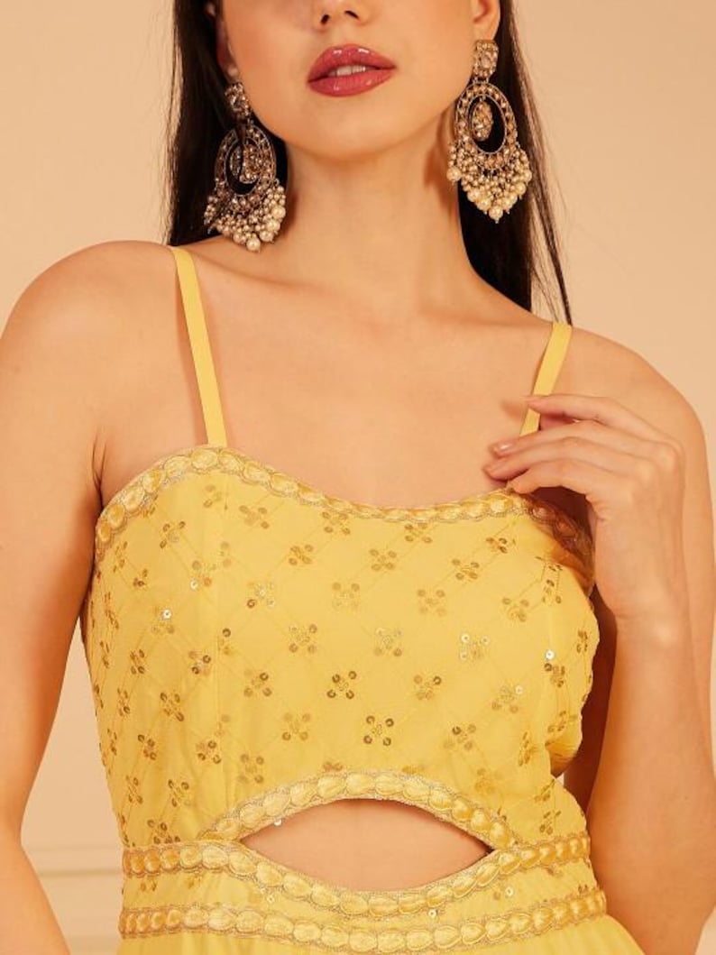 Pode incluir: Vestido de noite amarelo com decote em cora&ccedil;&atilde;o e recorte em forma de fechadura. O vestido &eacute; adornado com lantejoulas e um padr&atilde;o floral. A modelo usa brincos dourados grandes e ornamentados com p&eacute;rolas.