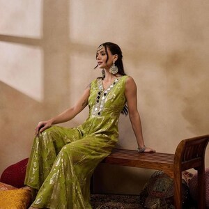 Op de afbeelding: Een vrouw draagt een groene jumpsuit met gouden borduurwerk en spiegelaccenten. Ze zit op een houten bank met bloemen op de voorgrond.