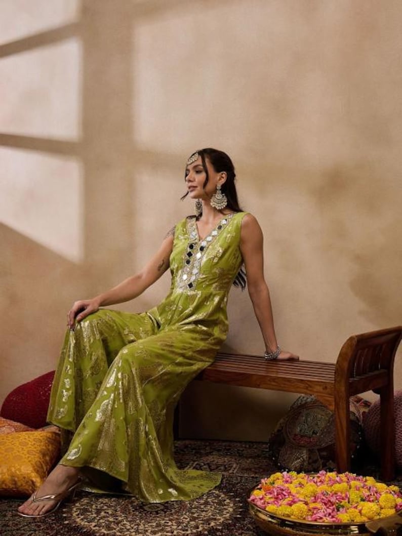 Op de afbeelding: Een vrouw draagt een groene jumpsuit met gouden borduurwerk en spiegelaccenten. Ze zit op een houten bank met bloemen op de voorgrond.