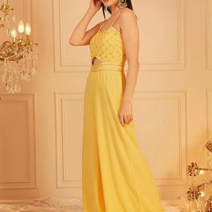 Pode incluir: Macac&atilde;o amarelo com al&ccedil;as finas e um detalhe vazado na cintura. O corpete &eacute; adornado com pequenos detalhes brilhantes. As cal&ccedil;as de pernas largas chegam at&eacute; o ch&atilde;o. A roupa &eacute; feita de tecido leve.