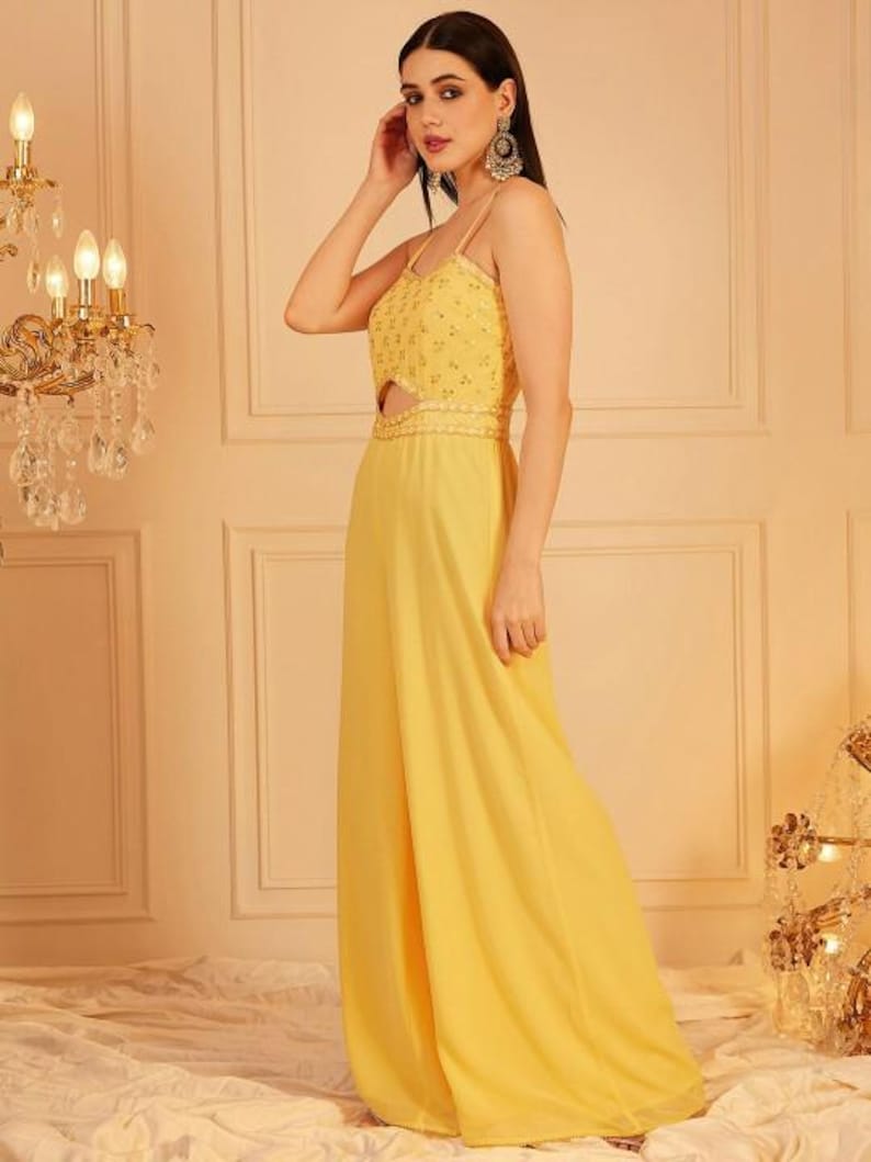 Pode incluir: Macac&atilde;o amarelo com al&ccedil;as finas e um detalhe vazado na cintura. O corpete &eacute; adornado com pequenos detalhes brilhantes. As cal&ccedil;as de pernas largas chegam at&eacute; o ch&atilde;o. A roupa &eacute; feita de tecido leve.