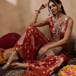 K&ouml;nnte beinhalten: Ein rotes und goldfarben besticktes Palazzo-Outfit mit V-Ausschnitt, verziert mit Spiegelakzenten. Das Outfit hat weite Hosenbeine und wird mit goldenen Sandalen getragen. Es ist ein traditionelles indisches Kleidungsst&uuml;ck.