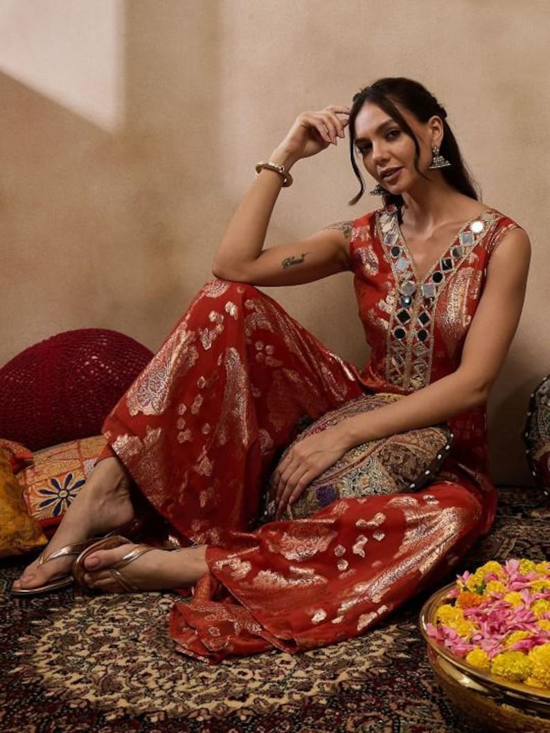 K&ouml;nnte beinhalten: Ein rotes und goldfarben besticktes Palazzo-Outfit mit V-Ausschnitt, verziert mit Spiegelakzenten. Das Outfit hat weite Hosenbeine und wird mit goldenen Sandalen getragen. Es ist ein traditionelles indisches Kleidungsst&uuml;ck.