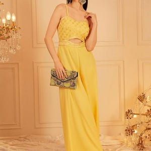 Pode incluir: Macac&atilde;o amarelo com recorte na cintura e al&ccedil;as finas. O corpete &eacute; adornado com lantejoulas. A pessoa segura uma clutch estampada. O fundo inclui um lustre e pain&eacute;is decorativos.