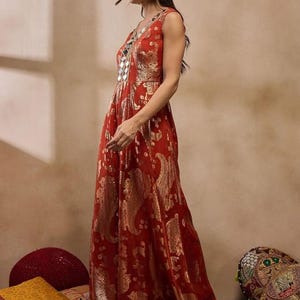 K&ouml;nnte beinhalten: Ein &auml;rmelloses, bodenlanges rotes Kleid mit goldenem Paisley-Muster. Das Kleid hat einen V-Ausschnitt mit Spiegelverzierungen. Das Model tr&auml;gt silberne Ohrringe und Sandalen.