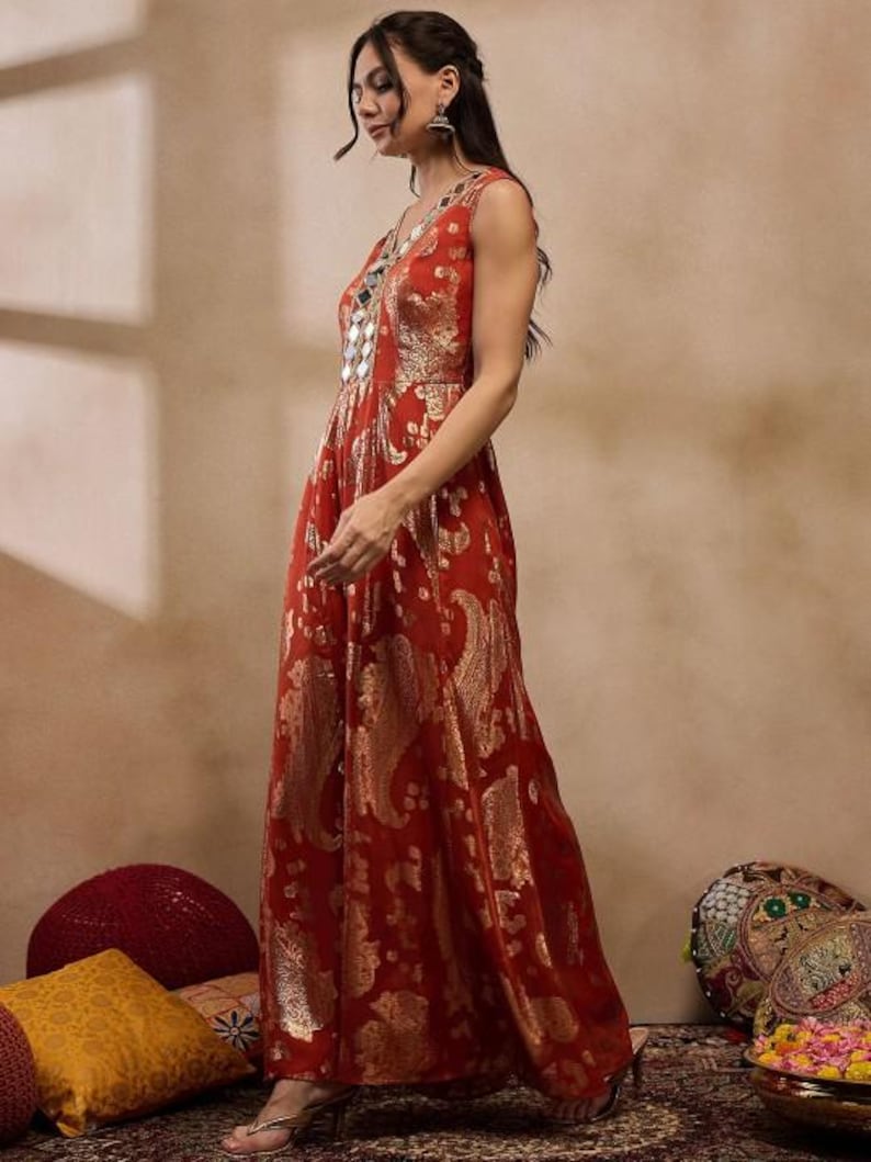 K&ouml;nnte beinhalten: Ein &auml;rmelloses, bodenlanges rotes Kleid mit goldenem Paisley-Muster. Das Kleid hat einen V-Ausschnitt mit Spiegelverzierungen. Das Model tr&auml;gt silberne Ohrringe und Sandalen.