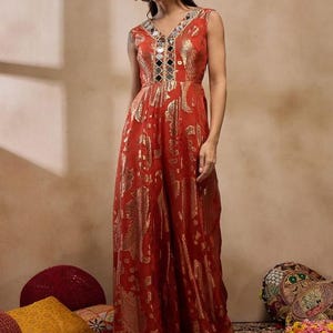 K&ouml;nnte beinhalten: Ein &auml;rmelloser, rostfarbener Jumpsuit mit V-Ausschnitt und weiten Beinen. Der Stoff hat ein goldfarbenes Metallic-Muster. Der Ausschnitt ist mit Spiegelakzenten verziert. Das Model tr&auml;gt silberne Ohrringe.