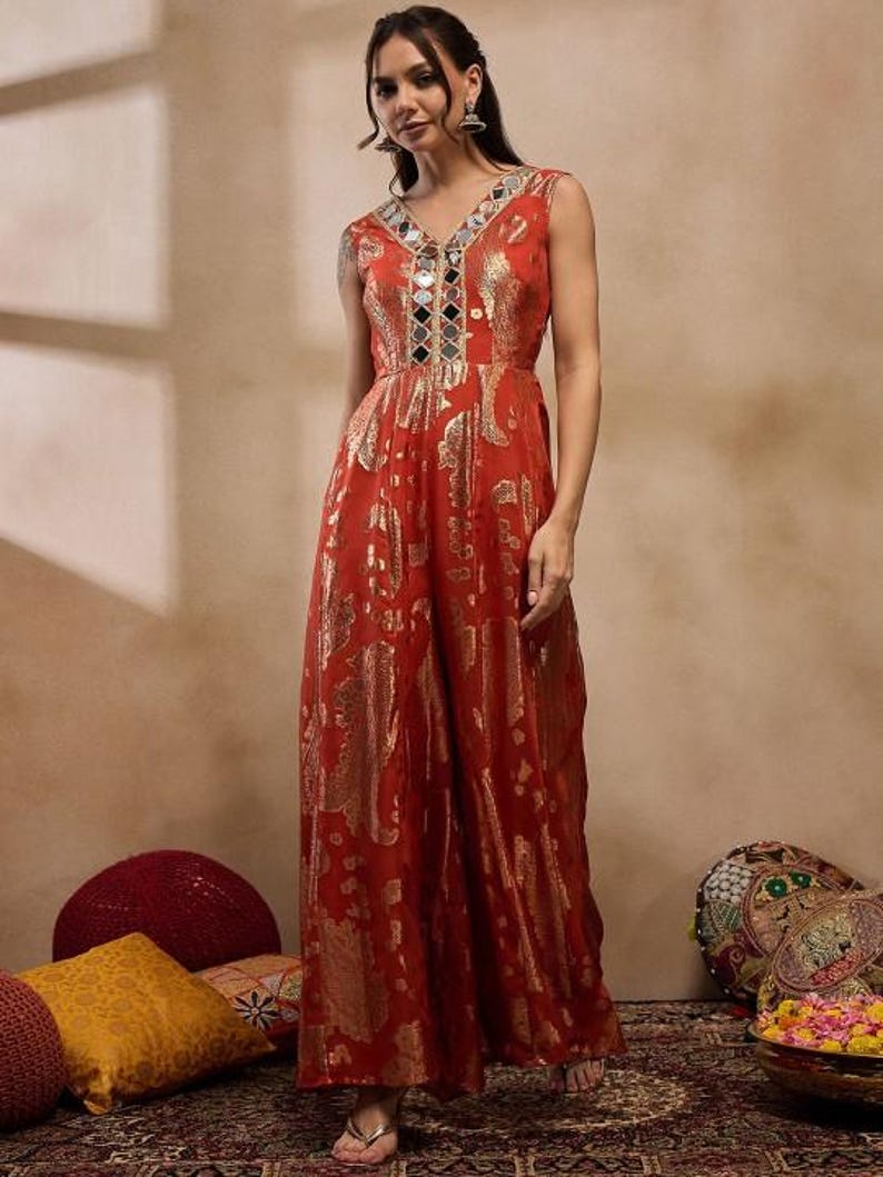 K&ouml;nnte beinhalten: Ein &auml;rmelloser, rostfarbener Jumpsuit mit V-Ausschnitt und weiten Beinen. Der Stoff hat ein goldfarbenes Metallic-Muster. Der Ausschnitt ist mit Spiegelakzenten verziert. Das Model tr&auml;gt silberne Ohrringe.