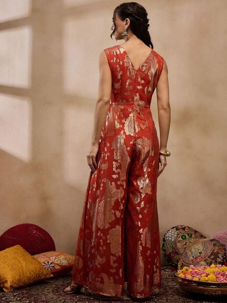 K&ouml;nnte beinhalten: Ein &auml;rmelloser, roter Jumpsuit mit V-Ausschnitt und weiten Beinen. Der Stoff hat ein goldenes Paisley-Muster. Das Model steht mit dem R&uuml;cken zur Kamera und pr&auml;sentiert das Design und die Passform des Kleidungsst&uuml;cks. Der Jumpsuit ist ein lebendiges Kleidungsst&uuml;ck.