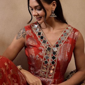 K&ouml;nnte beinhalten: Ein &auml;rmelloses rotes Kleid mit V-Ausschnitt, verziert mit kleinen Spiegeln und goldenen Verzierungen. Das Kleid hat ein Blumenmuster und wird mit silbernen Ohrringen und einem goldenen Armband getragen. Der Stil deutet auf einen festlichen oder formellen Anlass hin.
