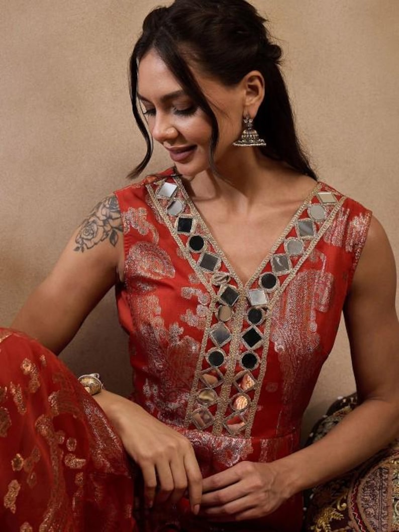 K&ouml;nnte beinhalten: Ein &auml;rmelloses rotes Kleid mit V-Ausschnitt, verziert mit kleinen Spiegeln und goldenen Verzierungen. Das Kleid hat ein Blumenmuster und wird mit silbernen Ohrringen und einem goldenen Armband getragen. Der Stil deutet auf einen festlichen oder formellen Anlass hin.