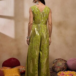 Op de afbeelding: Een groene jumpsuit met een gouden paisley print. De jumpsuit heeft een V-hals en is mouwloos. Het model draagt de jumpsuit en staat voor een beige achtergrond.
