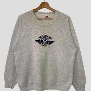 Könnte beinhalten: Ein grauer Pullover mit einem dunkelblauen gestickten Logo, auf dem "Dockers Since 1850 Levi's" steht. Das Logo zeigt einen geflügelten Anker und ein Seil.