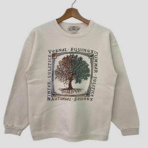 May include: A cream colored sweatshirt with a graphic of a tree with roots and leaves. The tree is surrounded by a square frame with the words "Vernal Equinox", "Summer Solstice", "Autumnal Equinox", and "Winter Solstice" written around it.