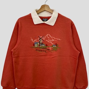 May include: Orange sweatshirt with a white collared shirt underneath. The sweatshirt has an embroidered image of a windmill and houses in a countryside setting. The text "Southern Lady" is on a black tag on the front of the shirt.
