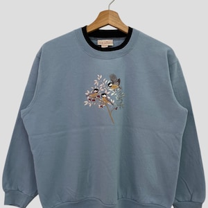 May include: A light blue sweatshirt with a black ribbed collar and embroidered birds on a branch. The sweatshirt has the text "Top Drawer" on the tag.