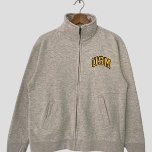 Könnte beinhalten: Ein grauer Reißverschluss-Pullover mit einem gelben gestickten Logo, auf dem "USM" steht.