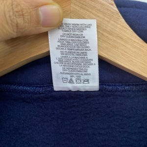 May include: White care label with instructions for washing and drying a navy blue sweatshirt. The label includes text in both English and Spanish. The text reads "Wash warm with like colors only non-chlorine bleach when needed tumble dry low do not iron or dry clean emblem lavado a maquina con aqua tibia con colores similares. Use solo blanqueador sin cloro secara temperatura baja no planche ni limpie en seco el emblema".