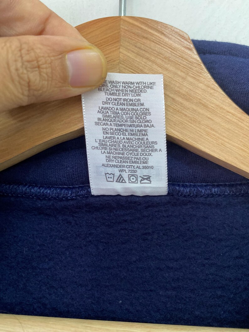 May include: White care label with instructions for washing and drying a navy blue sweatshirt. The label includes text in both English and Spanish. The text reads "Wash warm with like colors only non-chlorine bleach when needed tumble dry low do not iron or dry clean emblem lavado a maquina con aqua tibia con colores similares. Use solo blanqueador sin cloro secara temperatura baja no planche ni limpie en seco el emblema".