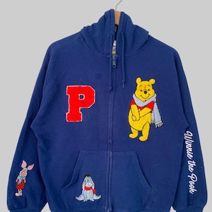 May include: A navy blue zip-up hoodie featuring a large red letter P, an embroidered Winnie the Pooh wearing a scarf, a small Eeyore graphic, and Piglet on the sleeve. The text Winnie the Pooh is printed in white on the other sleeve.