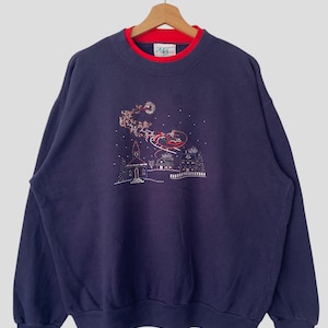 May include: A navy blue long-sleeve sweatshirt featuring a red inner collar and an embroidered Christmas scene on the chest. The design depicts a sleigh with reindeer flying over a small village with a church and house under a full moon.