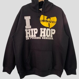 Puede incluir: Sudadera con capucha negra con un estampado gráfico blanco que dice "I ♥ HIP HOP IM FUCKING SERIOUS." El gráfico también incluye un logotipo amarillo de Wu-Tang Clan.
