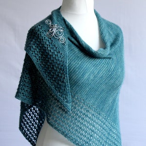 May include: A teal blue knitted shawl with a delicate openwork pattern. The shawl is draped over a white mannequin and features a silver metal pin.