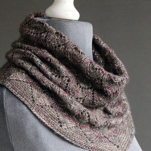 May include: A hand-knitted cowl scarf in shades of grey, brown, and pink, displayed on a mannequin. The scarf features a textured, openwork design with a triangular shape. The soft, warm accessory is perfect for cold weather.