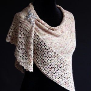 May include: A cream-coloured knitted shawl with an openwork pattern and a silver brooch. The shawl is displayed on a black mannequin, highlighting its texture and form. The shawl features a speckled design with touches of pink.