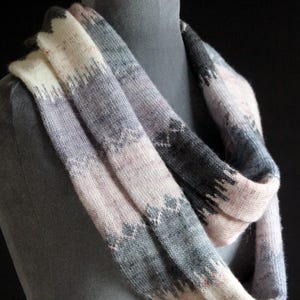 May include: A knitted infinity scarf in white, pink, and grey, displayed on a mannequin. The scarf has a geometric pattern and a soft, textured appearance, ideal for cold weather. The scarf is approximately 152 cm in circumference.