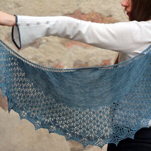 May include: A blue knitted shawl with a delicate lace pattern. The shawl is draped over the arm of a person wearing a white shirt.