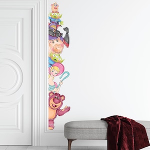 May include: A wall decal featuring characters from the Toy Story movie franchise. The characters are arranged vertically, with Woody, Buzz Lightyear, Jessie, and other characters visible. The decal is applied to a white wall next to a white door.