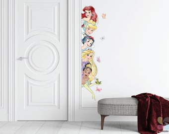Op de afbeelding: Een witte deur met een decoratief ontwerp staat naast een witte muur met een verticale lijn van Disney-prinsessen. De prinsessen zijn Ariel, Assepoester, Sneeuwwitje, Rapunzel en Tiana. Er zijn vlinders en een kikker op de muur.