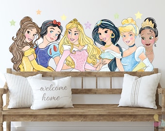 May include: A wall decal featuring six cartoon princesses, including Belle, Snow White, and Cinderella, with colorful star accents. The princesses are positioned above a wooden bench with two white pillows. One pillow has the text "welcome home".