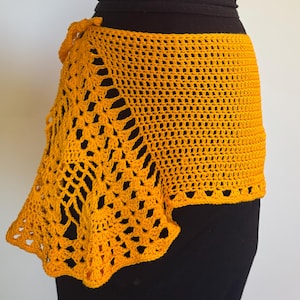 May include: Mustard yellow crochet skirt with a unique asymmetrical design. The skirt is handmade with a textured stitch pattern and a scalloped edge. The skirt is tied at the waist and is shown on a black mannequin.
