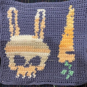 Könnte beinhalten: Gehäkeltes Quadrat mit einem Totenkopf- und Knochen-Design in Beige und Orange auf dunkelblauem Hintergrund. Eine Karotte mit grünen Blättern ist rechts auf dem Quadrat gehäkelt.