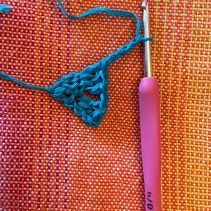 May include: A close-up of a turquoise crochet project in progress. A pink crochet hook with the text "5.0 mm" is visible in the image.