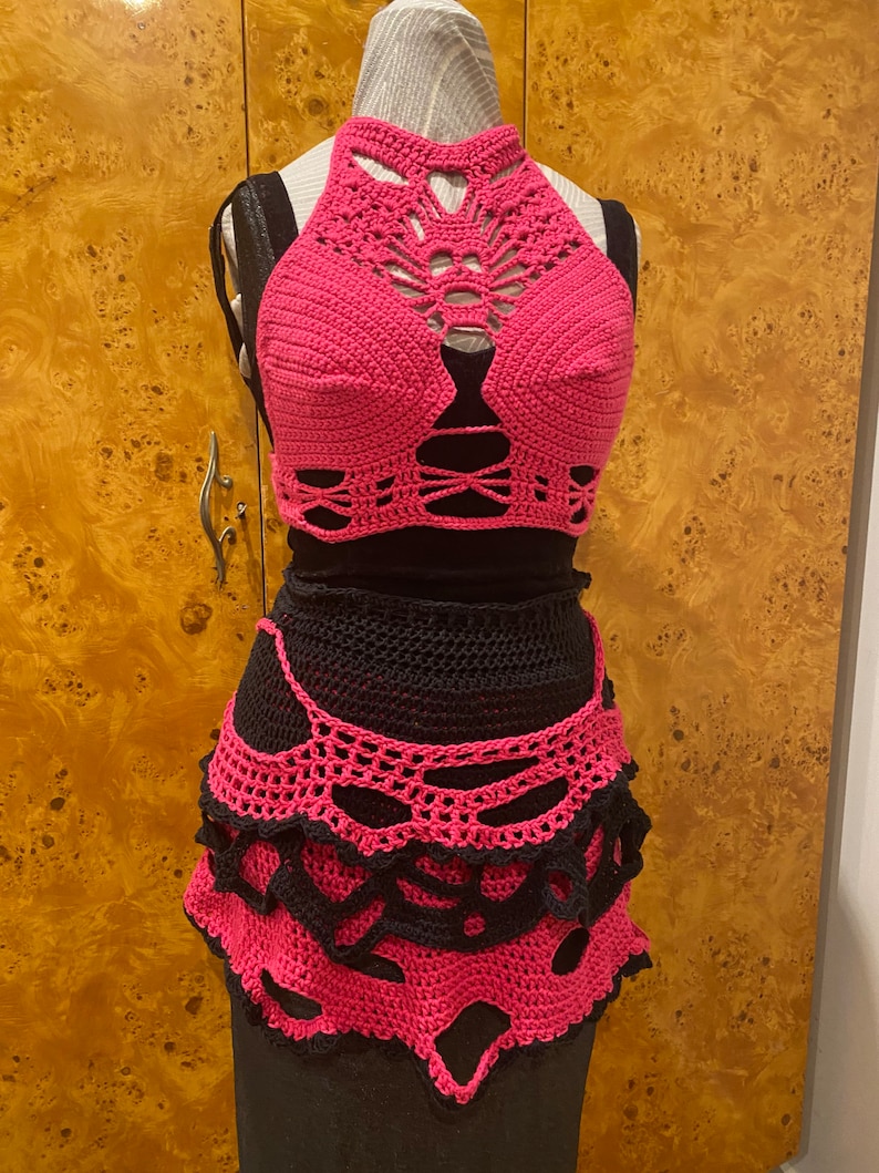 May include: A pink crocheted halter top and skirt set with a black mesh lining. The top has a delicate, intricate design with cutouts and the skirt has a layered, scalloped hem.